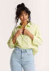 Limoengroene button-up shirt met een kraag, ontspannen pasvorm, lange mouwen opgerold; gecombineerd met lichtblauwe high-waisted jeans.