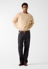 Beżowy sweter z dzianiny z prążkowanymi mankietami, zestawiony z ciemnymi jeansami denim. Brązowy skórzany pasek z okrągłą złotą klamrą, brązowe buty.