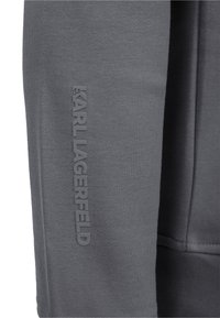 Gros plan sur un tissu gris avec un logo texte vertical en relief "KARL LAGERFELD" sur la couture de la manche d'un vêtement.