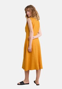 Mustard yellow linen dress with a fitted bodice and flared skirt, featuring cap sleeves and a knee-length design, paired with black slide sandals.