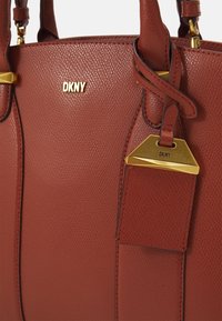 Sac à main en cuir rouge texturé avec ferrures dorées et logo embossé. Présente une forme structurée et une étiquette détachable inclinée.