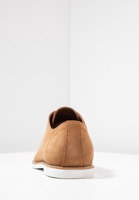 Chaussure en daim marron clair avec un bout arrondi et une semelle en caoutchouc blanc lisse. Présente des coutures à l'arrière et une finition légèrement texturée.