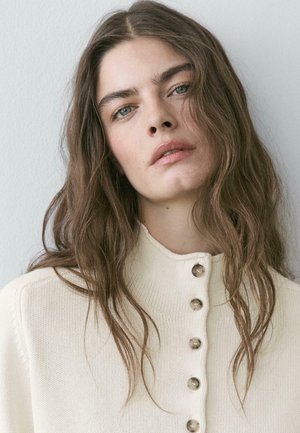 Kvinde med langt, bølget brunt hår iført en cremefarvet, knaplukket striktrøje, der ser let opad mod en ensartet lys baggrund.