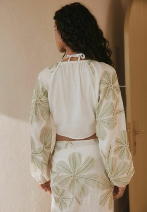 Sieviete ar cirtainiem matiem, tērpusies baltā divdaļīgā tērpā ar lielu zaļu ziedu izšuvumu, stāv telpās, skatoties prom no kameras.