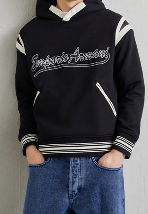 Person trägt schwarzen Hoodie mit weißen Streifen und Schriftzug „Emporio Armani“, kombiniert mit blauen Jeans, Hände in den Taschen, stehend.