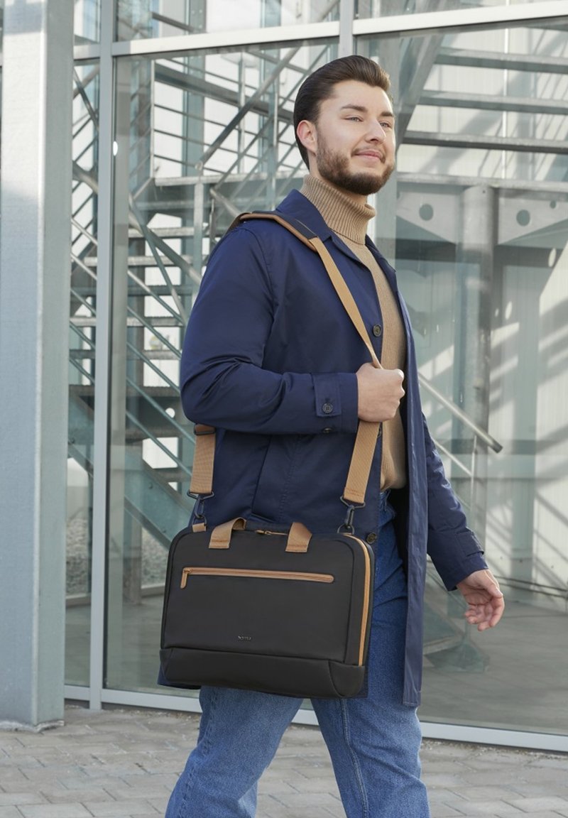 Mann, der im Freien geht, trägt einen blauen Mantel und einen beigen Rollkragenpullover, hält eine schwarz-beige Umhängetasche und befindet sich in der Nähe eines gläsernen Gebäudes mit sichtbarer Metalltreppe.