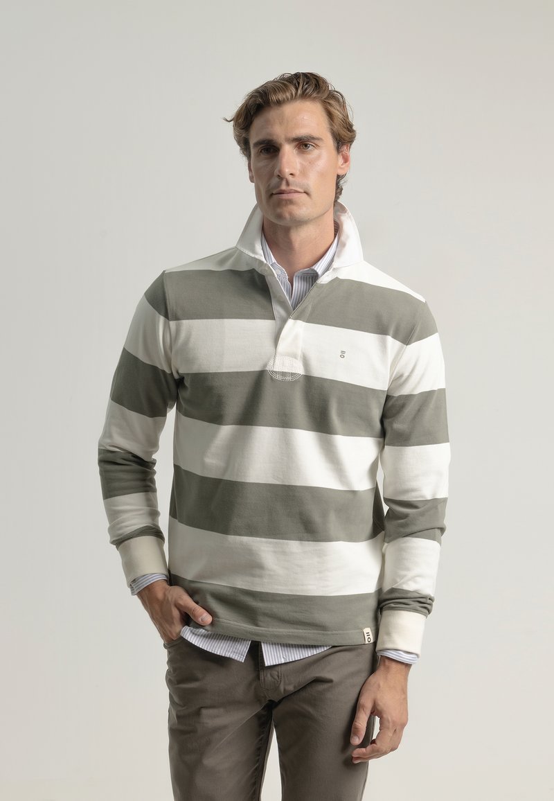 Camisa de rugby a rayas en verde y blanco, con collar, mangas largas, un pequeño logo y un aspecto superpuesto sobre una camisa a rayas azul.