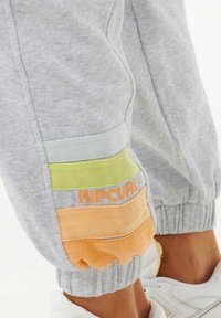 Pantalones de chándal grises con un diseño en bloques de color y rayas horizontales en azul claro, verde y naranja. Incluye puños elásticos y la marca "RIPCURL".