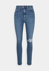Jeans skinny de cintura alta en denim azul oscuro, con una rodilla desgastada, diseño de cinco bolsillos y cierre de botón metálico.