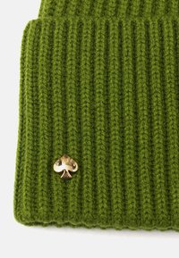 Tejido de punto acanalado verde con un pequeño emblema de pala dorada brillante adjunto cerca del borde inferior.