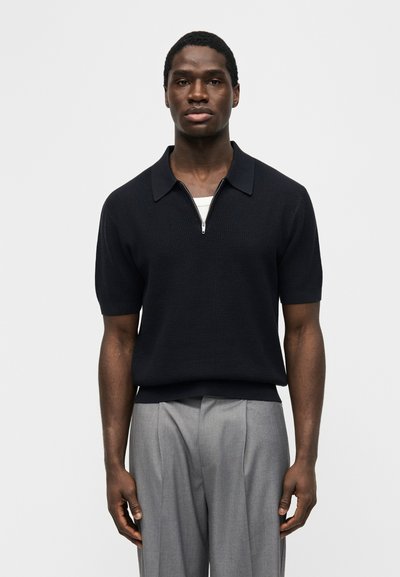 Νέος Μαύρος άνδρας που φοράει μαύρο πλεκτό πόλο με κοντά μανίκια και φερμουάρ, καθώς και ανοιχτό γκρι πλισέ παντελόνι, πόζαρε μπροστά σε απλό φόντο.