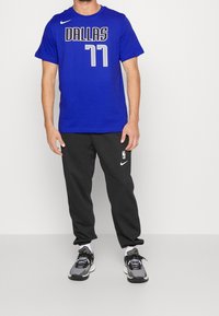 Námořnická modrá Nike tričko s nápisem "DALLAS" bílou barvou a "77" stříbrnou. Černé tepláky a šedé tenisky dokončují celý outfit.