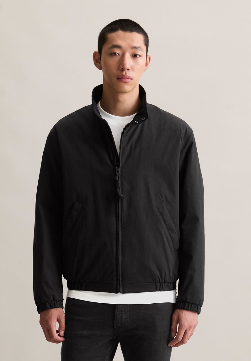 Jonge man in een zwarte zip-up jack over een wit shirt en zwarte broek, staand tegen een effen lichte achtergrond, met het gezicht naar voren.