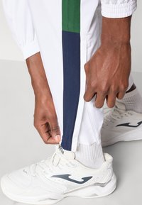 Calças desportivas brancas com uma faixa lateral em azul escuro e verde, apresentando punhos elásticos e detalhes de fecho na bainha. Usadas com ténis brancos.