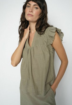 Žena s vlnitými hnedými vlasmi, oblečená v svetlo olivovom riasenom tielku bez rukávov a v zodpovedajúcich nohaviciach, s rukou dotýkajúcou sa krku.