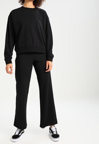 Sweat-shirt noir à coupe décontractée, associé à un pantalon noir à jambes larges. Le mannequin porte des baskets noires avec des accents blancs. Texture de tissu lisse.