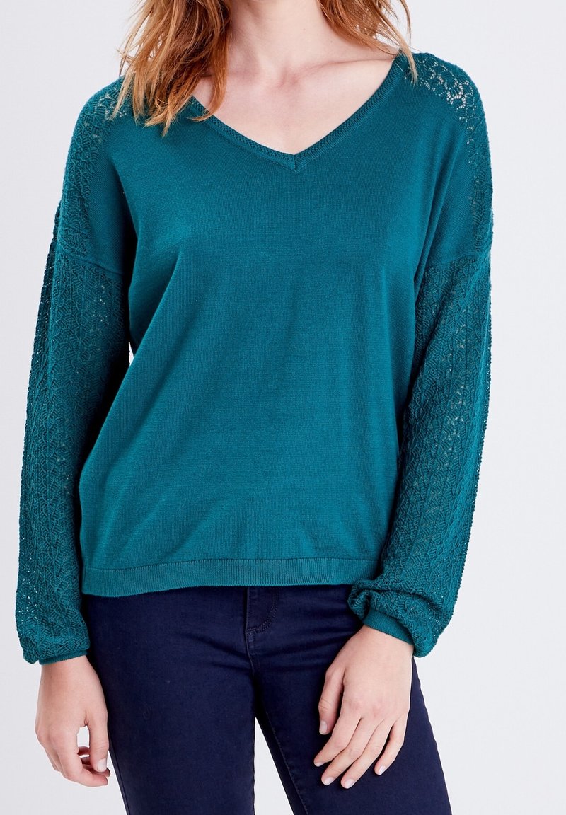 Femme portant un pull col en V couleur sarcelle avec des manches en dentelle tricotée, associé à un pantalon bleu foncé, debout devant un fond uni.