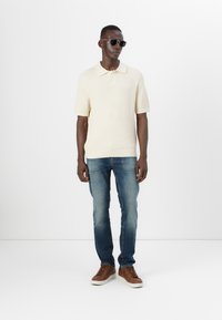 Polo shirt crem cu dungă, cu mâneci scurte, asortat cu blugi blue slim-fit și teniși din piele maro. Modelul poartă ochelari de soare negri.