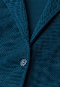 Gros plan d'un blazer bleu texturé avec un revers cranté et une fermeture à un seul bouton avec du texte autour du bord du bouton.