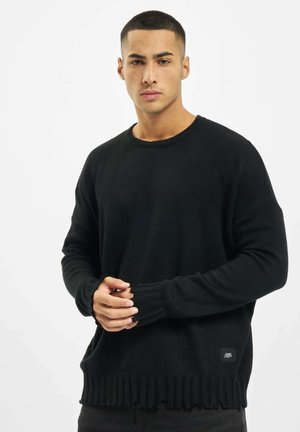 Black knitted sweater with a round neckline, textured fabric, and fringe detail at the hem. Ribbed cuffs and a logo patch on the bottom.