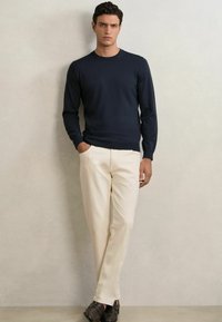Maglione a maglia blu navy con maniche lunghe, abbinato a pantaloni beige e scarpe marrone scuro. Il maglione ha un collo rotondo e polsini aderenti.