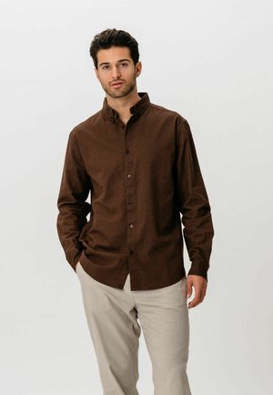 Mężczyzna ubrany w ciemnobrązową koszulę na guziki i jasnobeżowe spodnie, stojący z jedną ręką w kieszeni na jednolitym tle.