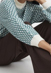 Pull en maille bleu sarcelle avec motif moucheté blanc, doté d'un col et de poignets côtelés crème. Associé à un pantalon large marron foncé.