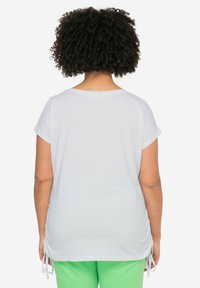 Top blanco de manga corta con un corte holgado, que presenta cordones laterales y una textura suave, combinado con pantalones verdes.