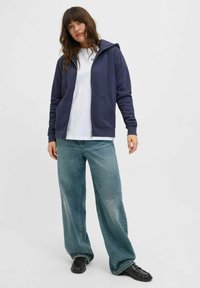 Felpa con zip color navy, con lacci, tasche anteriori, indossata sopra una maglietta bianca e jeans larghi di un blu chiaro, completata da scarpe nere.