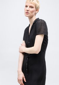 Γυναίκα με κοντά ξανθά μαλλιά που φοράει μαύρο φόρεμα με κουμπιά και κοντά μανίκια, στέκεται μπροστά σε απλό ανοιχτό φόντο.