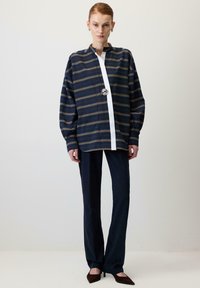 Stribet skjorte med knapper i marineblå og guld, med en hvid frontknaplukning og en rund sølvspænde, kombineret med marineblå bukser og mørke hæle.