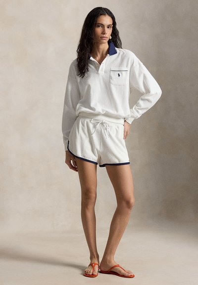 Nő áll, kezét a csípőjén tartja, fehér hosszú ujjú pólót, hozzá illő fehér rövidnadrágot sötétkék szegéllyel, és narancssárga papucsot visel semleges háttér előtt.