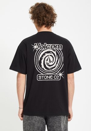 Person med krøllet hår iført en sort Volcom t-shirt med et hvidt hvirvlende galaksedesign og teksten "STONE CO." på ryggen.
