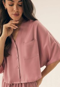 Różowa, jedwabna bluzka z guzikami, z dekoltem w serek, krótkimi szerokimi rękawami i kieszonką z przodu. Gładka faktura i luźny krój.
