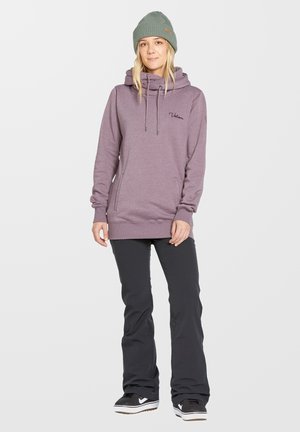 Lichtpaarse hoodie met kangoeroezakken, een trekkoord capuchon en geborduurd logo. Gecombineerd met zwarte broek en een groene gebreide beanie.