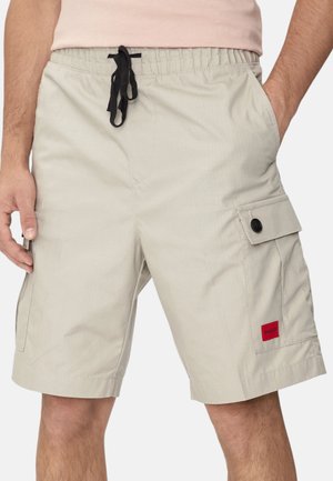 Uomo che indossa pantaloncini cargo beige con tasche laterali e coulisse nera, mostrato dalla vita al ginocchio, con le mani vicino alle tasche.