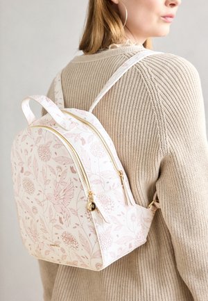 Femme portant un pull beige et un sac à dos blanc avec un motif floral rose et un colibri, ainsi que des détails de fermeture éclair dorés.