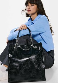 Borsa nera lucida con manici a catena argentati, caratterizzata da una texture liscia e una forma rettangolare strutturata. Posizionata di fronte a un modello seduto.