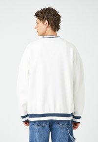 Sudadera blanca de corte relajado, con puños y dobladillo de canalé en rayas azul marino y blancas, que presenta un clásico cuello redondo y una textura suave.