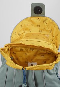 Sac à dos vert clair avec un intérieur jaune orné de motifs sur le thème de l'extérieur. Comprend une poche transparente et une fermeture à bouton rond.