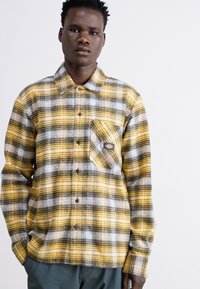 Camicia flanella a quadri gialli e neri con abbottonatura frontale, maniche lunghe e taschino sul petto con un patch logo. Tessuto testurizzato.
