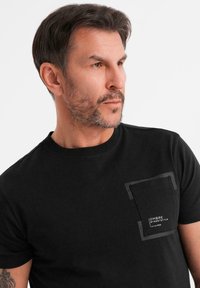 Svart bomull T-shirt med rund hals, med en ficka med rektangulär design och vit text på. Slät yta, korta ärmar.