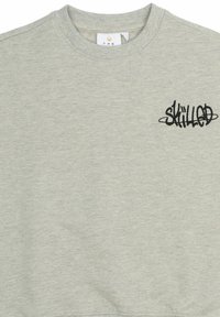 Sivi pamučni dukserica s okruglim izrezom i crnim izvezenim logotipom "SKILLED" na lijevoj strani prsnog koša. Glatka tekstura i ležeran dizajn.
