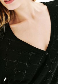 Cardigan noir avec un décolleté en V profond, une texture douce et un motif en grille avec de petites touches scintillantes. Deux boutons visibles à l'avant.