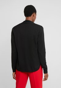 Schwarzer langärmliger Cardigan mit geripptem Saum und Kragen, in lockerer Passform und weicher Textur, kombiniert mit roten Hosen.