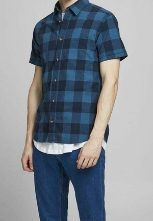 Hombre vistiendo una camisa de botones de manga corta a cuadros azul y negra sobre una camiseta blanca, con jeans azules, de pie frente a un fondo sencillo.