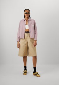 Ljusrosa rutigt knäppskjorta, vit croptop, beige vida shorts, guldskor med svarta snören och svarta strumpor; solglasögon bärs.