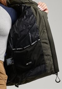 Oliven grøn jakke med en sort, tekstureret foring indvendig. Har et Superdry-logo bånd, lynlåse og lommer. Viser indre konstruktionsdetaljer.