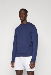 Navyblauwe sweatshirt van een zachte, textuur-achtige stof met een ronde hals en lange mouwen. Bevat een klein wit logo op de borst. Witte shorts.