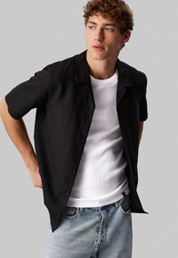 Camisa negra de manga corta con botones y un corte relajado, hecha de tejido ligero, llevada sobre una camiseta de tirantes blanca con texturas en relieve, combinada con jeans de color azul claro.